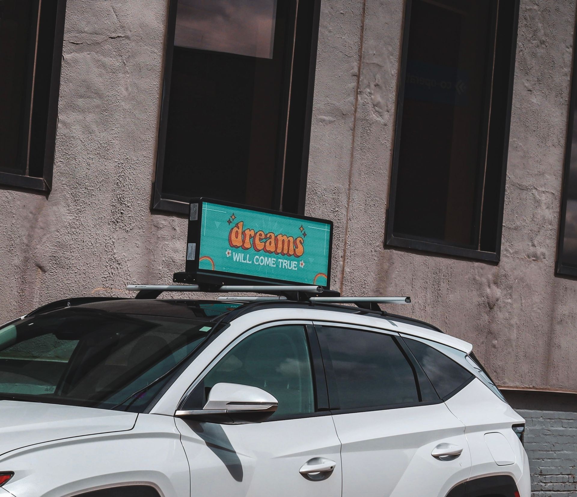 White SUV with rooftop digital sign reading 'dreams will come true' parked by a building.